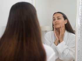 Uma mulher, de roupão de banho, pratica a rotina de beleza de cuidados com a pele em casa, estudando-se no espelho do banheiro Por demopicture