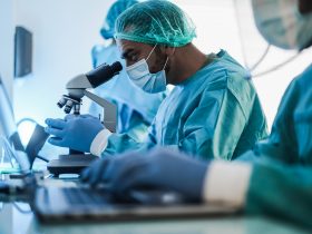 Cientista médico em traje de materiais perigosos trabalhando com microscópio e laptop dentro do laboratório Por DisobeyArtPh