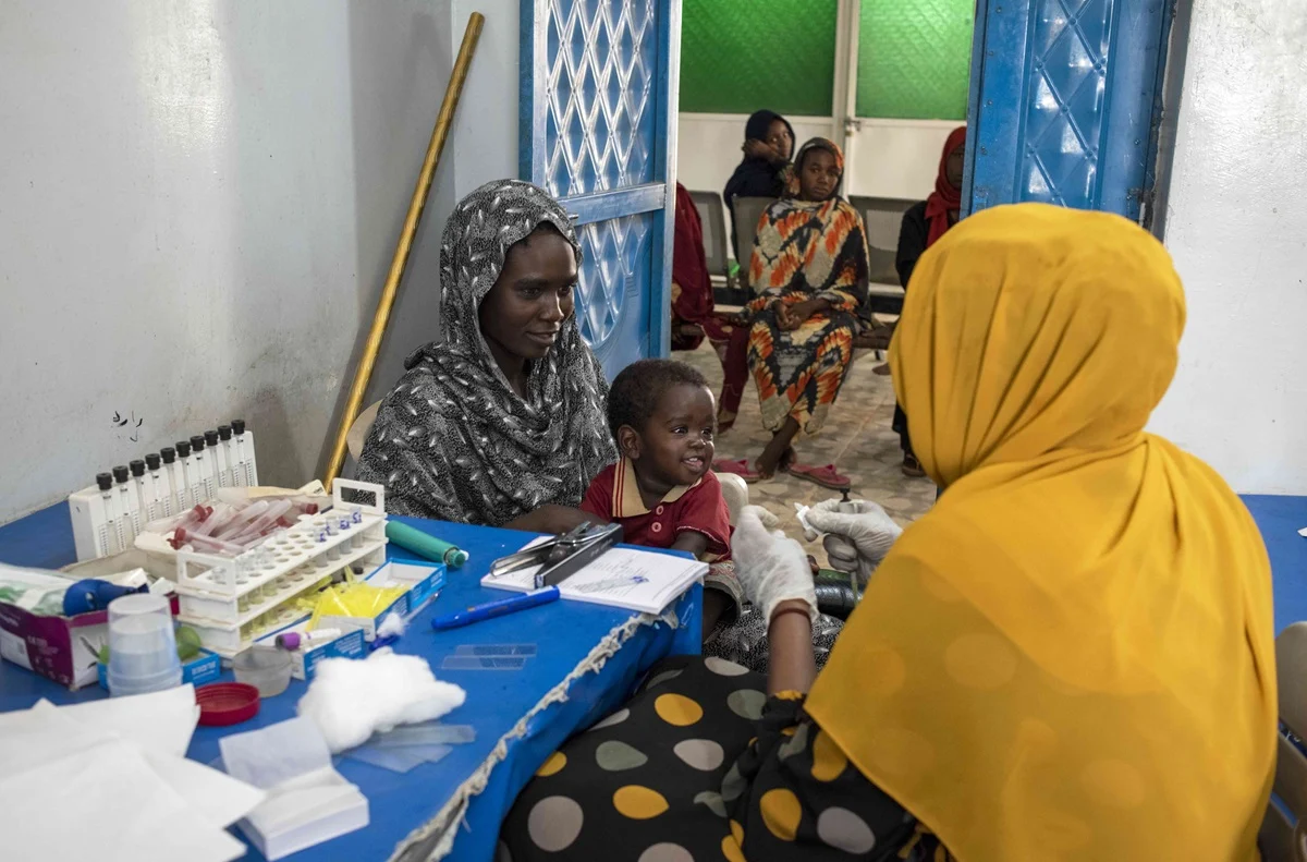 Uma mãe leva seu filho para um exame de sangue em uma unidade de saúde para mulheres e crianças no campo de IDP Abu Shouk, em Darfur do Norte. Em 2003, um conflito eclodiu em Darfur e forçou milhões de pessoas a fugir de suas casas | Foto de Lindsay Mackenzie para OMS